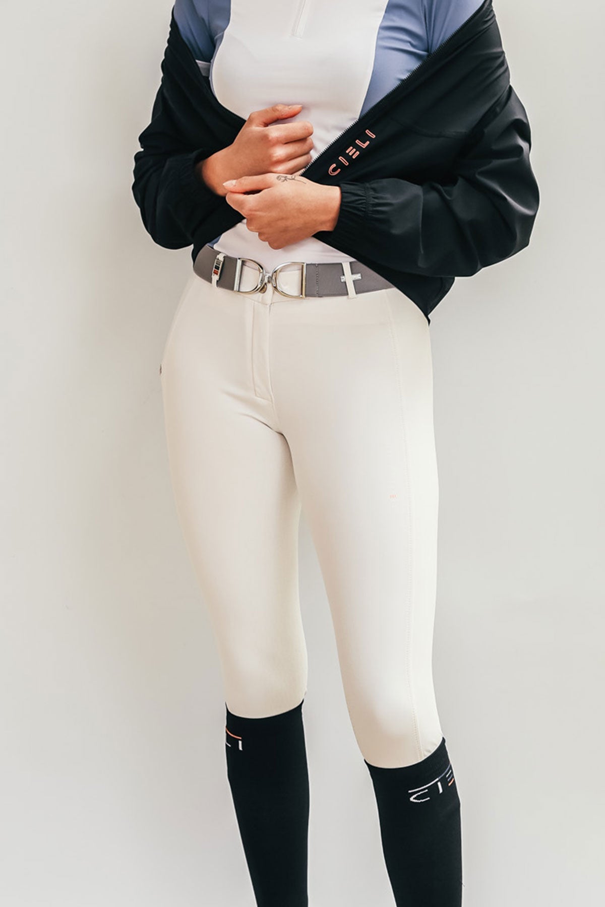 Person wearing white riding pants with a black jacket and black socks on a plain background