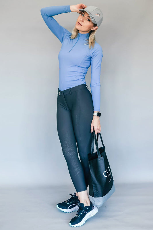 Woman wearing a blue top and grey riding breeches with a black bag, standing against a plain background