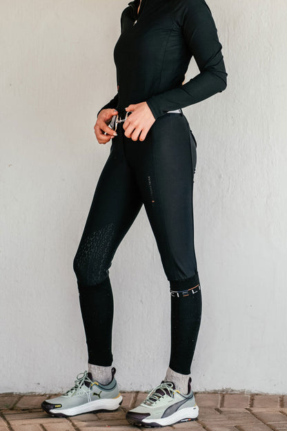 Person wearing black athletic breeches and shoes against a white wall.