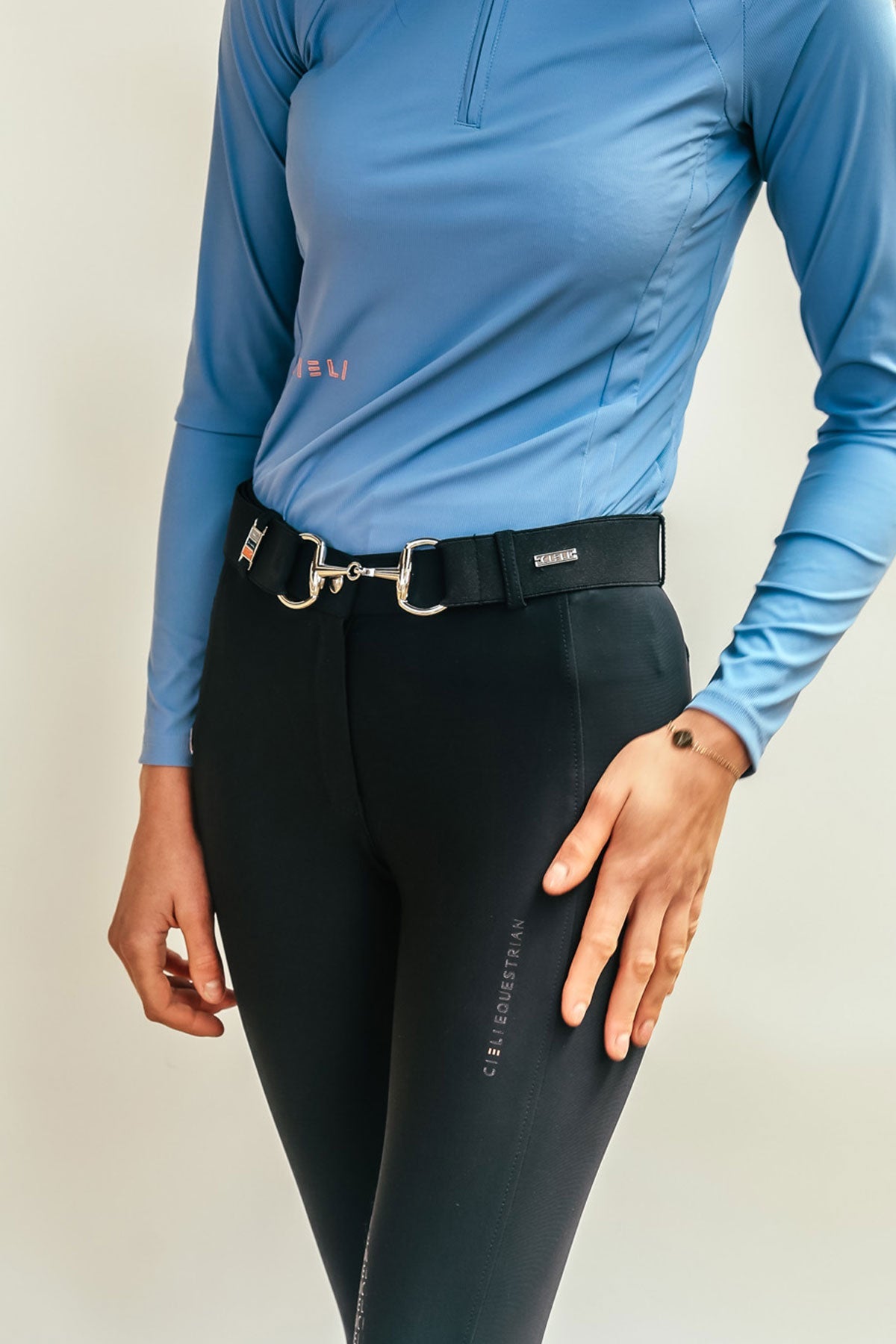 Person wearing a blue long-sleeve top and black riding pants on a plain background