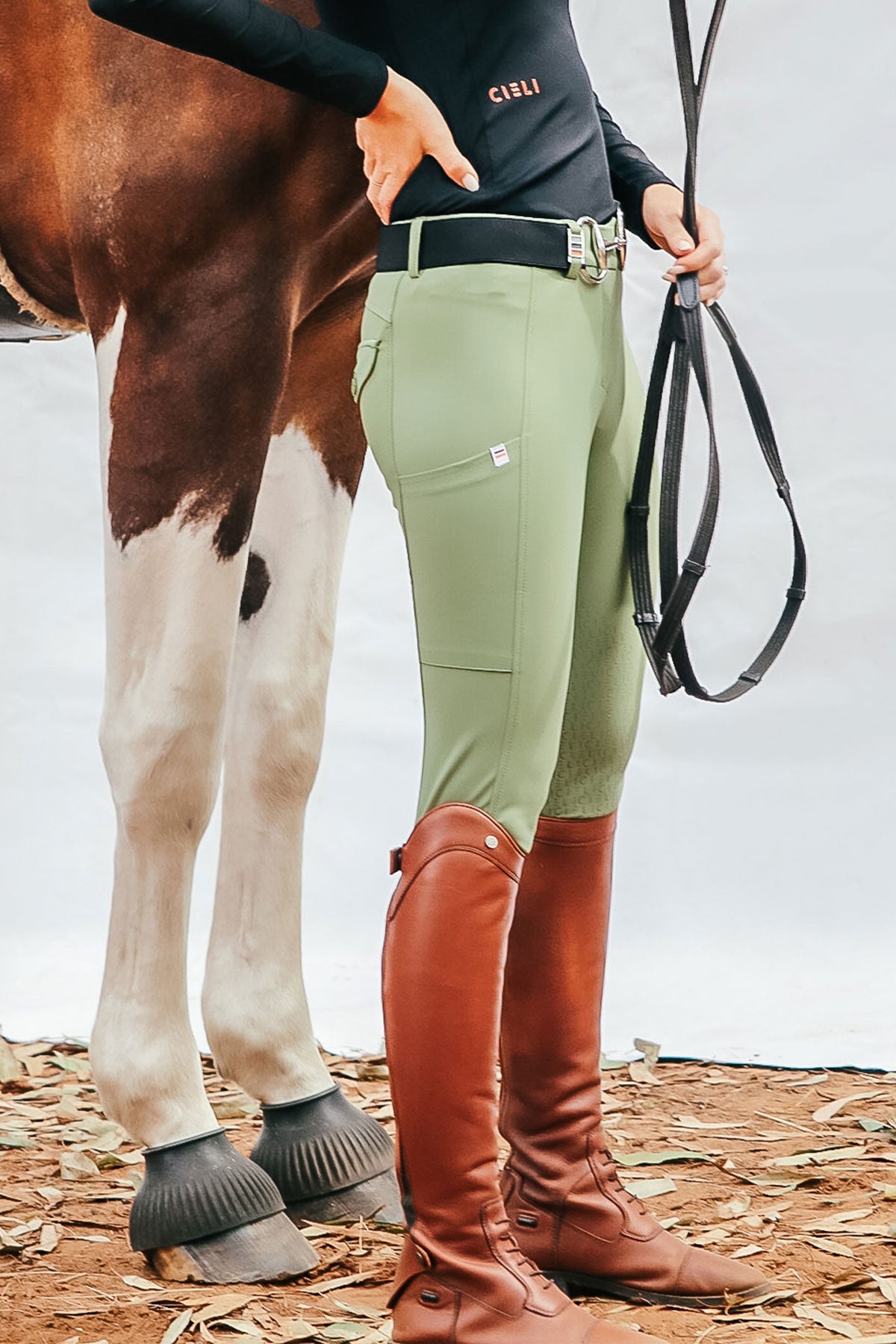 Person wearing green riding pants and brown riding boots standing next to a horse.