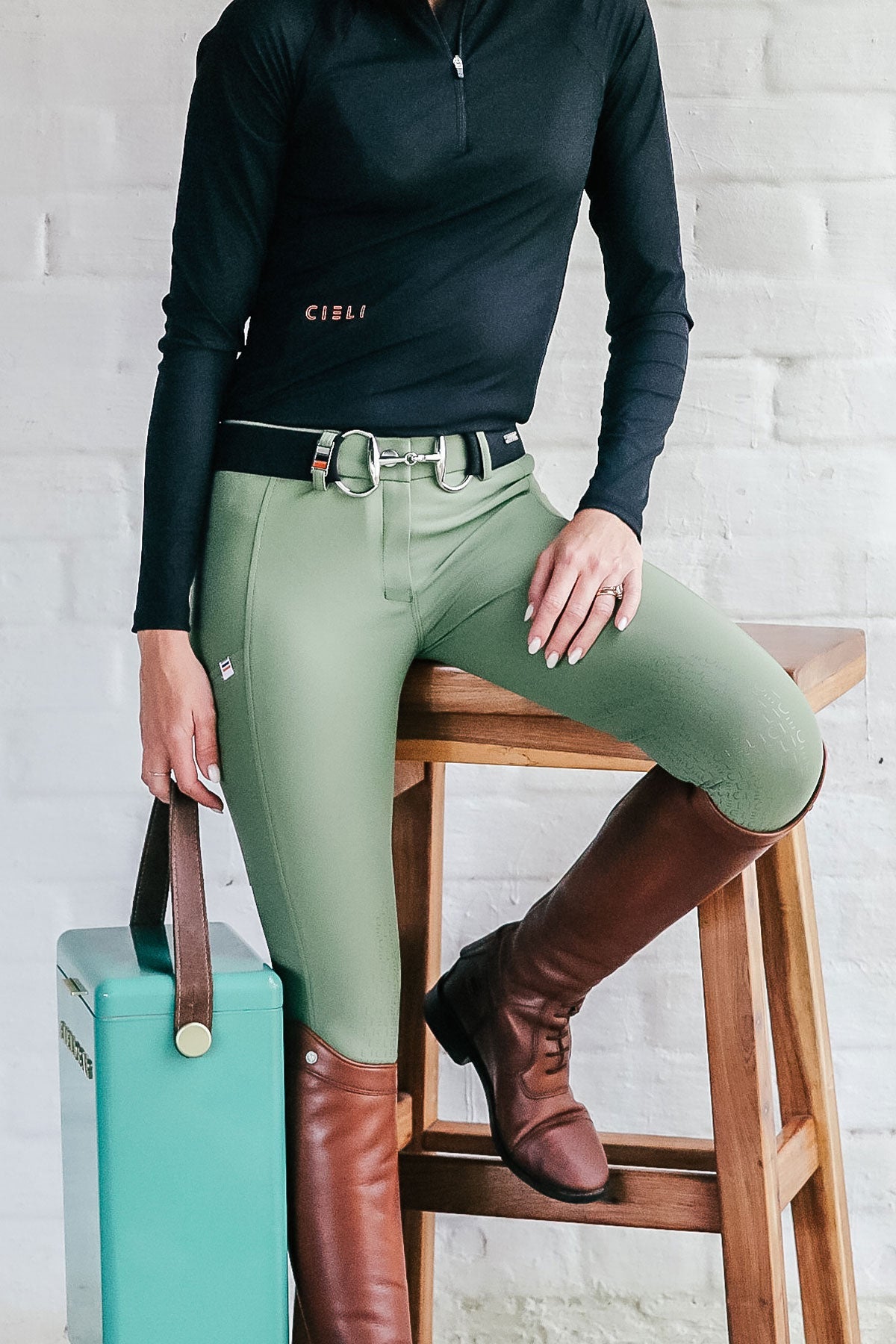 Person wearing green riding pants and brown boots, sitting on a wooden stool with a white brick wall background.