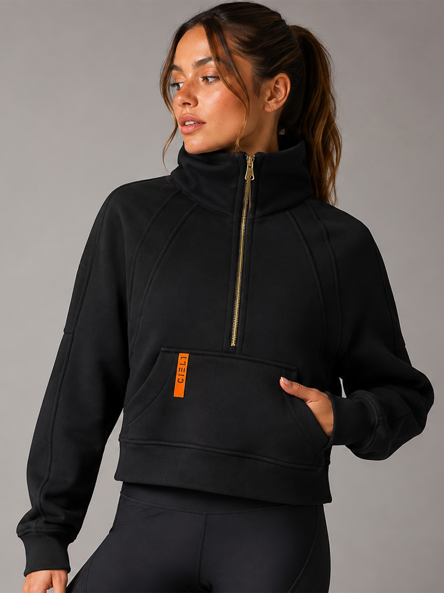 A woman wearing a black cropped half-zip sweatshirt with a high collar, gold zipper, and a small orange vertical logo tab on the pocket, paired with black leggings against a gray studio background.