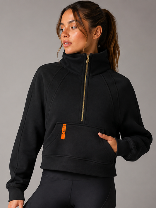 A woman wearing a black cropped half-zip sweatshirt with a high collar, gold zipper, and a small orange vertical logo tab on the pocket, paired with black leggings against a gray studio background.