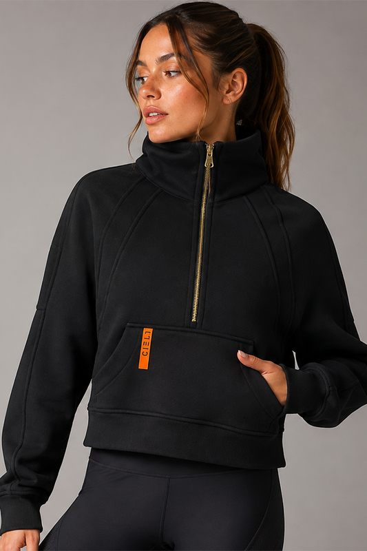 A woman wearing a black cropped half-zip sweatshirt with a high collar and a small orange vertical logo tab on the pocket, paired with black leggings, against a plain gray studio background.