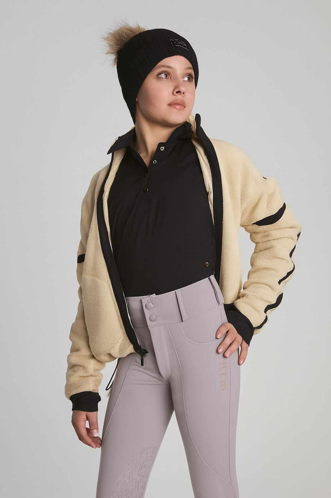 Person wearing a beige jacket with black accents, grey equestrian riding pants on a plain background