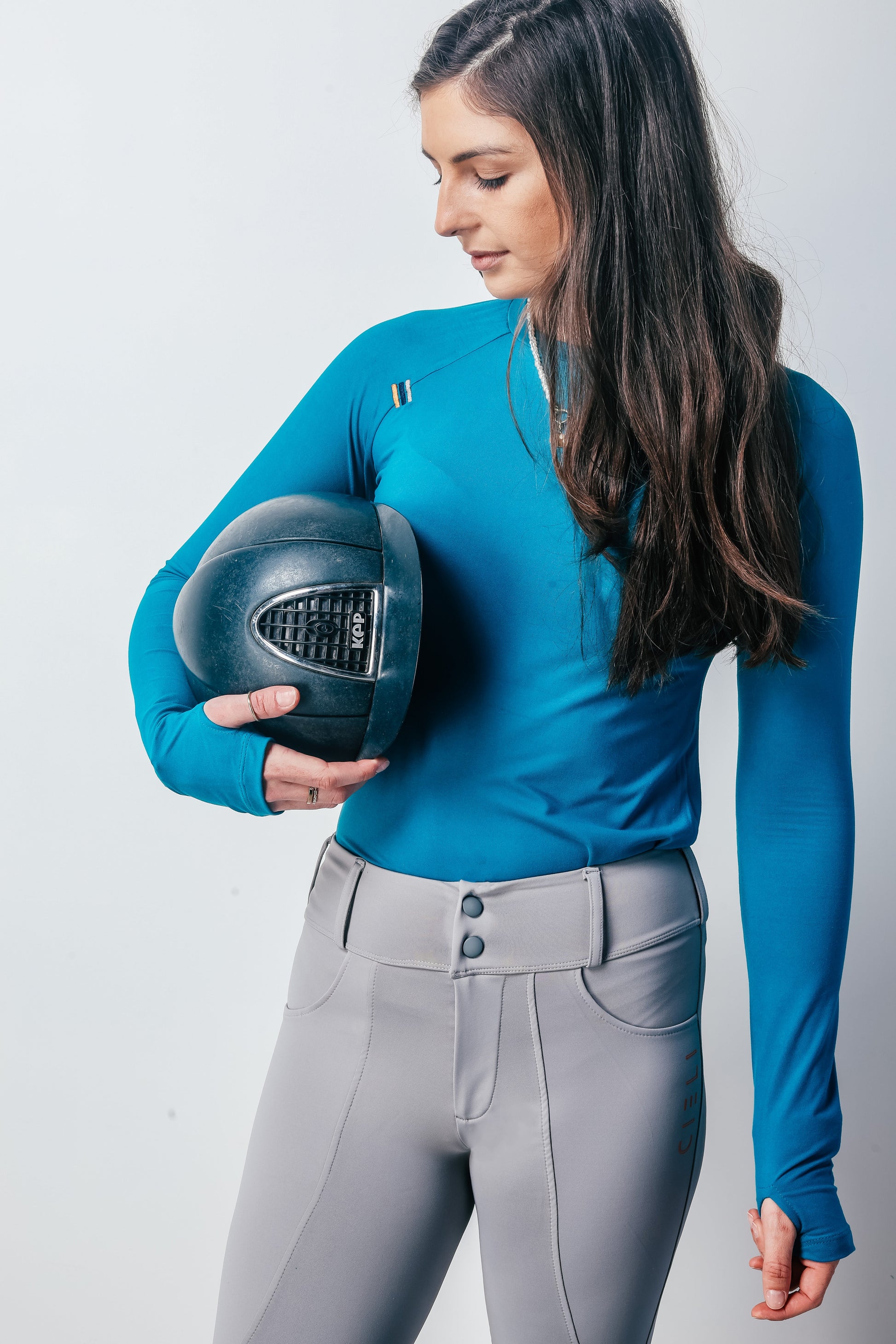 Woman in equestrian attire holding a helmet against a plain background
