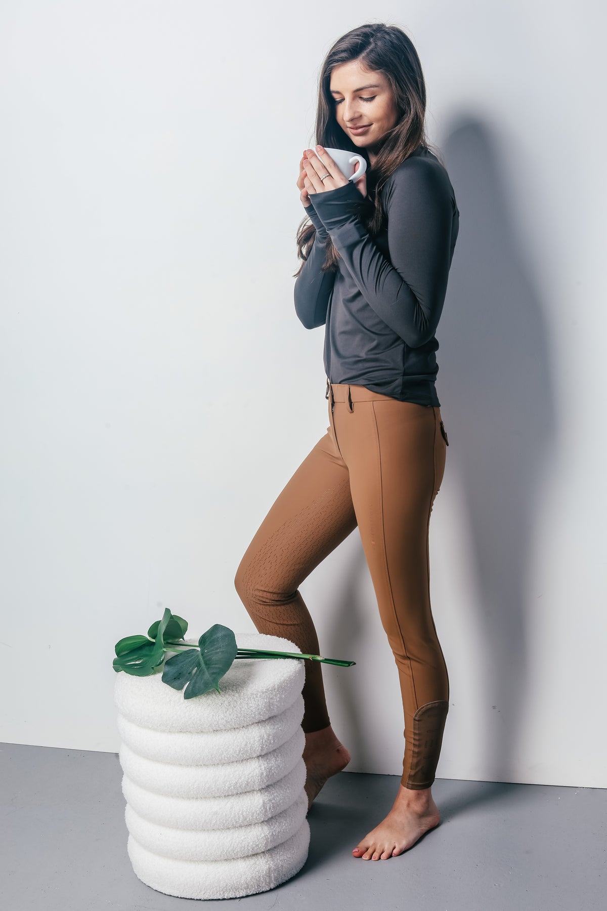 A woman stands barefoot against a light gray wall, wearing a dark gray long-sleeve top and brown leggings, holding a white mug. A white cylindrical stool with green leaves sits on the floor beside her.