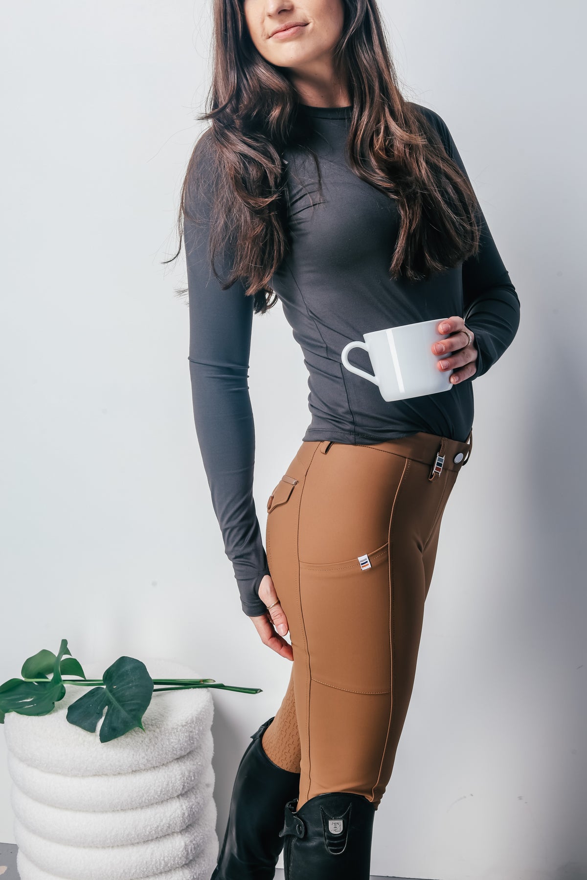 A woman modeling a dark gray long-sleeve athletic top and brown riding breeches, holding a white mug. She is standing next to a white textured stool with green leaves against a light gray background.
