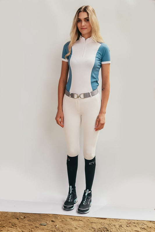 A woman modeling Berlin Performance Breeches by Cieli Equestrian. The breeches are white with orange accents, featuring a cell phone pocket with a logo label, belt loop with a pendant, and a subtle silicone print on the left leg.